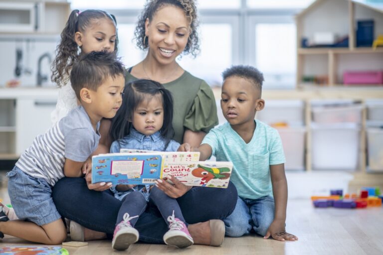 A preschool teacher sits on the floor in her classroom. Her students sit on and around her while she reads them a story. They are all engaged and look at the book.